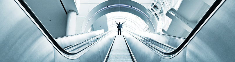 A man raises his arms in a celebration of success as he stands at the top of an escalator in a modern office building.