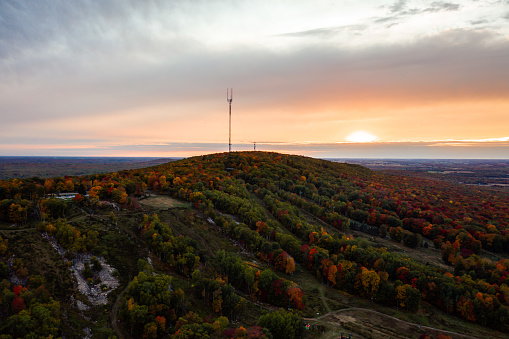 Beautiful aerial drone photograph of rib mountain with colorful fall leaves or autumn foliage lining the grassy ski runs at sunset as the cloudy sky turns orange and pink above the horizon.