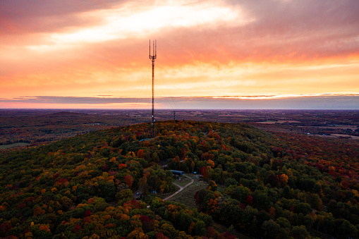 Beautiful aerial drone photograph of rib mountain as the sky erupts with orange and pink colored clouds beyond the broadcast tower at sunset with colorful fall leaves or autumn foliage on hill below.