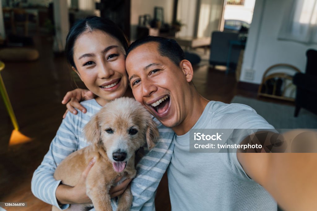 Junge erwachsene asiatische Paar hält einen Welpen, der ein Selfie von einem Telefon mit Home Interior im Hintergrund. 30er Jahre reifen Mann und Frau mit Hund Haustier, die eine Familie Fotoaufnahmen. - Glückliches Gruppenporträt. - Lizenzfrei Familie Stock-Foto