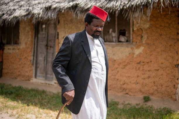 An African Older Man in Red Muslim Taqiyyah Fez Hat And Blazer Walking with a stick for limping people Near the Basic Hut with Thatched roof in Small Remote Village in Tanzania, Pemba island, Zanzibar Archipelago. un hombre mayor africano en el negro musulmán taqiyyah fez sombrero y blazer caminando con un palo para cojear personas cerca de la cabaña básica con techo de paja en pequeña aldea remota en tanzania, isla de pemba, archipiélago de zanzíbar