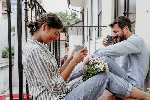 Cute and fun guy with a beard and a girl sitting on the balcony having their coffee and big laugh together. Both being casually dressed while she’s holding a flowers between her legs.