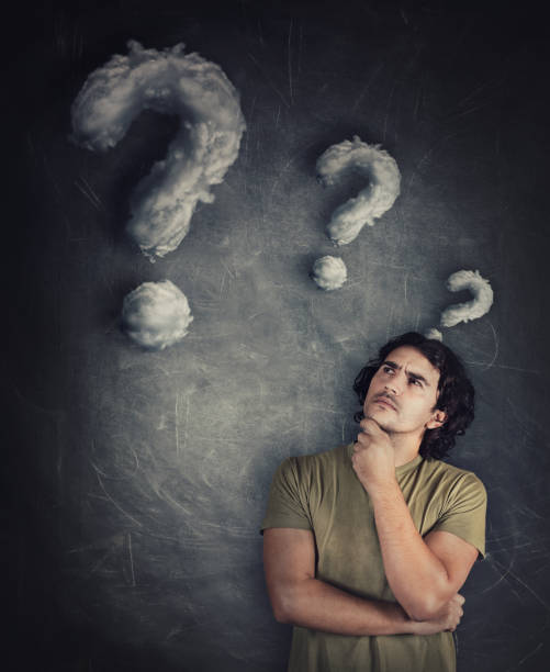 istock Confused young man, curly hair, keeps hand under chin, thoughtful gesture, looking suspicious aside as cloud shaped interrogation marks leaving his head. Puzzled guy, has many questions, quiz concept. 1286383520