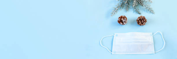 istock Protective medical mask, spruce branches and pine cone on blue background. Christmas and New Year during pandemic of coronavirus infection. Flat lay, top view, copy space. Banner. 1286383898