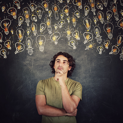 Pensive young man, long curly hair, keeps hand under chin, thoughtful gesture, standing behind a blackboard with different lightbulbs sketches. Positive thinking for multiple ideas. Genius concept.
