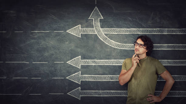 istock Pensive young man stands over chalkboard background with multiple straight arrows sketches, one of them changes direction bending by upwards trajectory. Different thinking concept, breaking the rules 1286387737