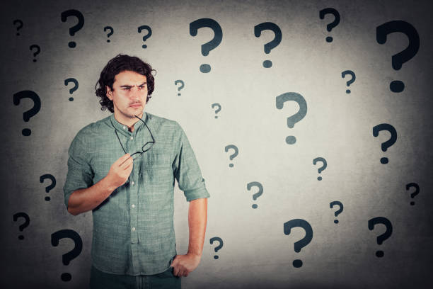 istock Puzzled man holds eyeglasses in his hand, thoughtful gesture, looking suspicious aside. Confused guy, thinking expression, different interrogation marks surrounds him. Unsure teen thinking for answer 1286387997
