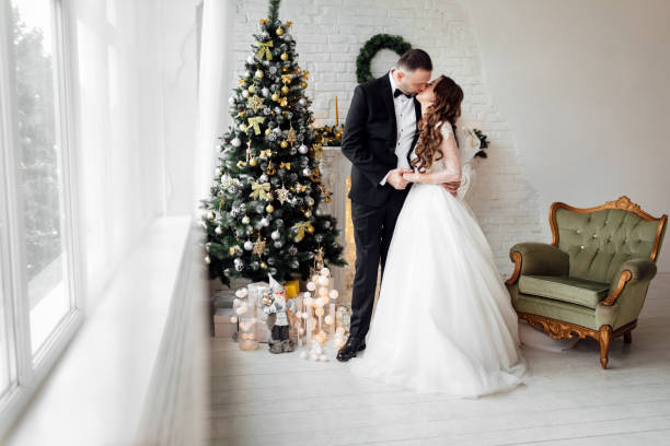 jeunes couples dans la mariée et le marié d’amour posant dans le studio sur le fond décoré avec l’arbre de noël dans leur jour de mariage à noël. profitez d’un moment de bonheur et d’amour. - christmas wedding bride winter photos et images de collection