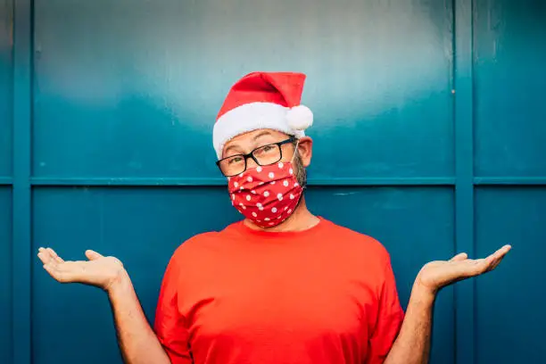 Photo of Portrait of cheerful and desolated adult man with red santa hat and face mask for coronavirus covid-19 virus emergency - concept of christmas and holidays alone during lockdown