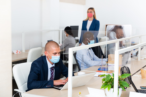 Portrait of confident manager wearing medical mask working on laptop in open plan office. Concept of precautions and social distancing in coronavirus pandemic