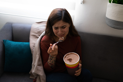 Depressed and heartbroken young latin woman eating chocolate ice cream while crying on the sofa. Hispanic woman feeling lonely