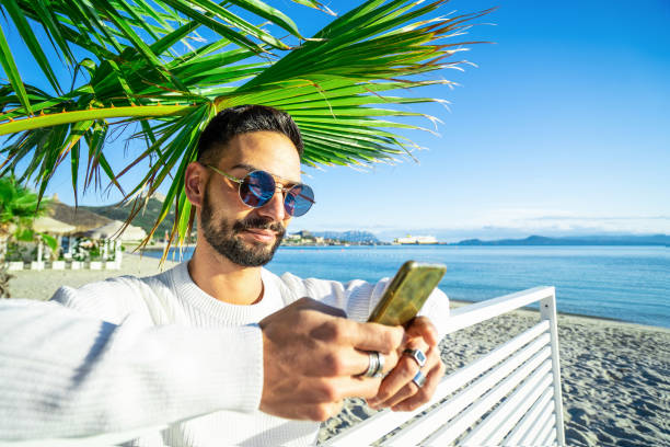 istock Handsome Caucasian bearded man with round sunglasses using smartphone under a palm on the beach - Stylish male person texting on phone messaging his friends and sharing his alone winter sea vacation 1286443831