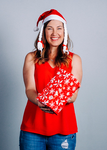 Portrait of smiling mature woman holding Christmas present. Happy female is wearing Santa hat. She is standing against gray background.