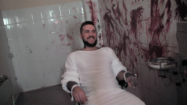 Crazy man in straitjacket tied to a chair, killer psychopath in a room smiles with bloodied walls overdose Crazy man in straitjacket tied to a chair, killer psychopath in a room with bloodied walls. drug addiction tattoos stock pictures, royalty-free photos & images