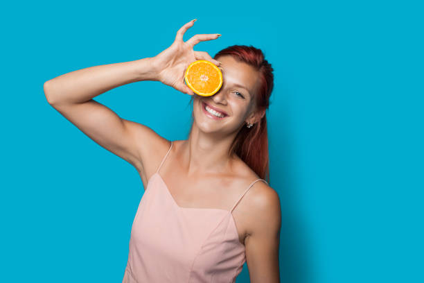 Red haired woman is covering her eye with a sliced lemon posing on a blue studio wall Red haired woman is covering her eye with a sliced lemon posing on a blue studio wall woman applying wallcovering stock pictures, royalty-free photos & images