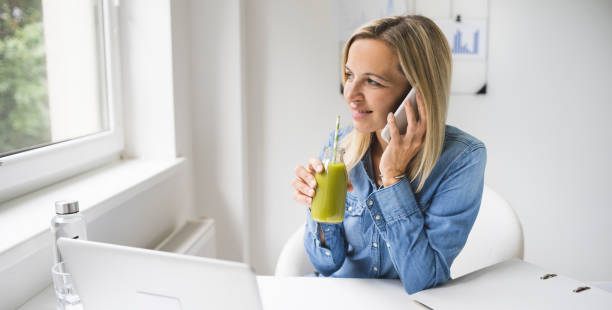 donna che beve frullato detox sano in ufficio davanti al laptop mentre parla al telefono - blueberry smoothie milk shake drink foto e immagini stock