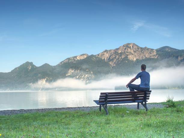 istock Man Sit Lone On A Bench In Park Next Lake. Mountain Range With Amazing View Over Smooth Lake 1286464847