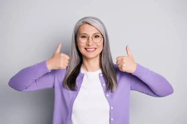 Photo of charming old mature lady white hair raise two hands show thumb-up, shiny smile wear eyewear white shirt violet cardigan isolated grey color background