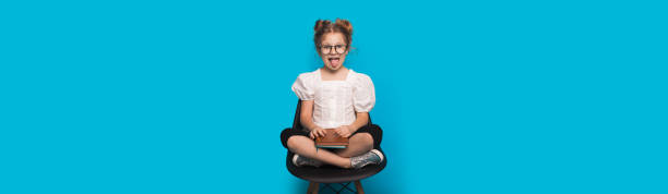 caucasian girl with glasses is smiling and showing tongue while sitting in armchair holding a book on a blue studio wall - preschooler childhood outdoors cheerful imagens e fotografias de stock