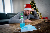 Conceptual image of hand sanitizer, face mask and out of Focus background of depressed man home alone in self isolation at christmas. COVID-19 Lockdown and quarantine on holidays and mental health.