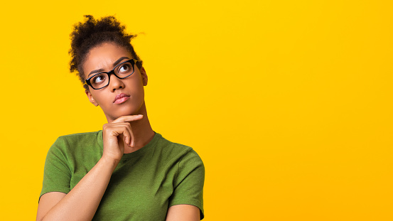 Making Decision. Closeup headshot of thoughtful african american lady in spectacles thinking about question, touching chin and looking away at free space isolated over yellow studio background, banner