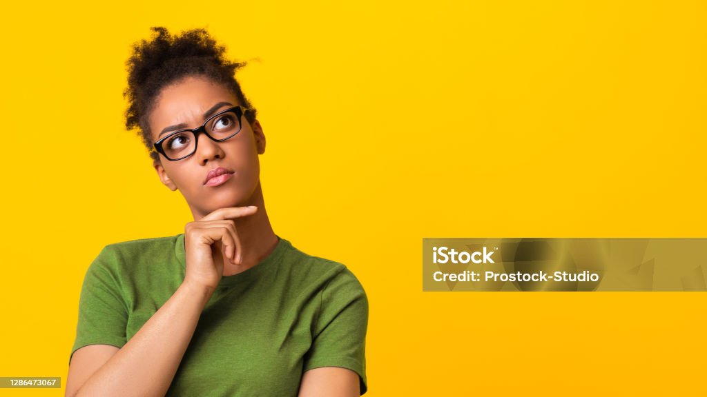 Black lady in eyeglasses thinking about question, touching chin Making Decision. Closeup headshot of thoughtful african american lady in spectacles thinking about question, touching chin and looking away at free space isolated over yellow studio background, banner Asking Stock Photo