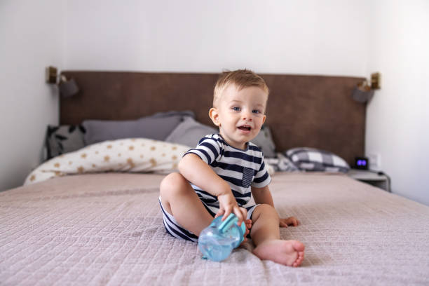 istock Cute little blond boy smiling and looking at camera with his beautiful blue eyes while sitting on bed in bedroom and holding his bottle with water. 1286485573
