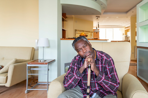 An old sad African-American woman with a cane is sitting in an armchair in the living room at home.