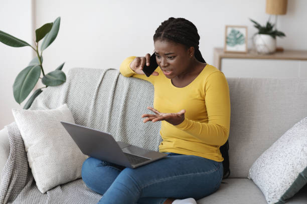 istock Frustrated black lady using laptop and talking on cellphone at home 1286488986
