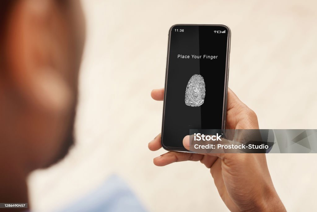 Man holding phone with fingerprint scanning app Biometric Identification Concept. Over The Shoulder View Of Black Man Holding Mobile Phone In Hand, Showing App For Fingerprint Scanning With A Zone To Touch With Thumbprint Icon On The Device Screen Fingerprint Stock Photo