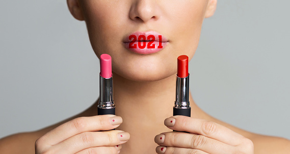 2021 Makeup Beauty Trends. Woman Choosing Lipstick Holding Cosmetics Near Face, With New Year Number On Lips Posing Over Gray Background. Upcoming Year And Make-Up Trend Concept. Panorama, Cropped