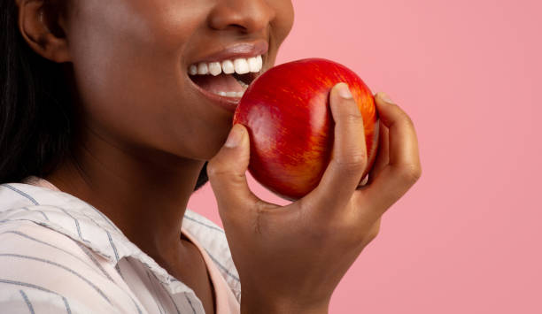 Dental care. Cropped view of black lady with healthy teeth eating ripe red apple over pink background, copy space Dental care concept. Cropped view of millennial black lady with healthy teeth eating ripe red apple over pink background, copy space. Banner design. Healthy nutrition for beautiful smile apple bite stock pictures, royalty-free photos & images