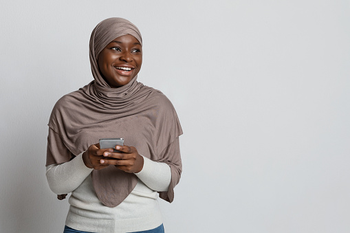 Mobile Offer. Cheerful Black Muslim Woman In Hijab Holding Smartphone And Looking Away At Copy Space, Smiling Lady Standing On Light Studio Background Using Cellphone, Enjoying Modern Communication