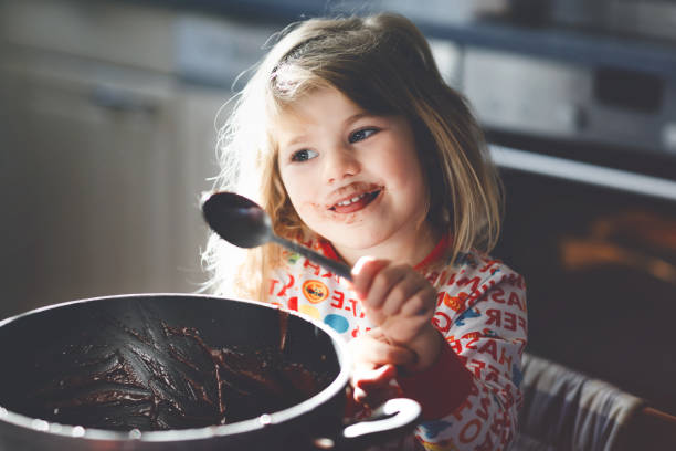 Cute toddler girl eating chocolate dough rests with spoon and fingers from pot. Happy child licking sweet dough for muffins or cake, helping in home kitchen, indoors. Cute toddler girl eating chocolate dough rests with spoon and fingers from pot. Happy child licking sweet dough for muffins or cake, helping in home kitchen, indoors chocolate fingers stock pictures, royalty-free photos & images