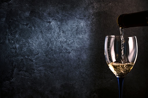 A glass with white wine pouring from a bottle on a dark gray background. Copy space