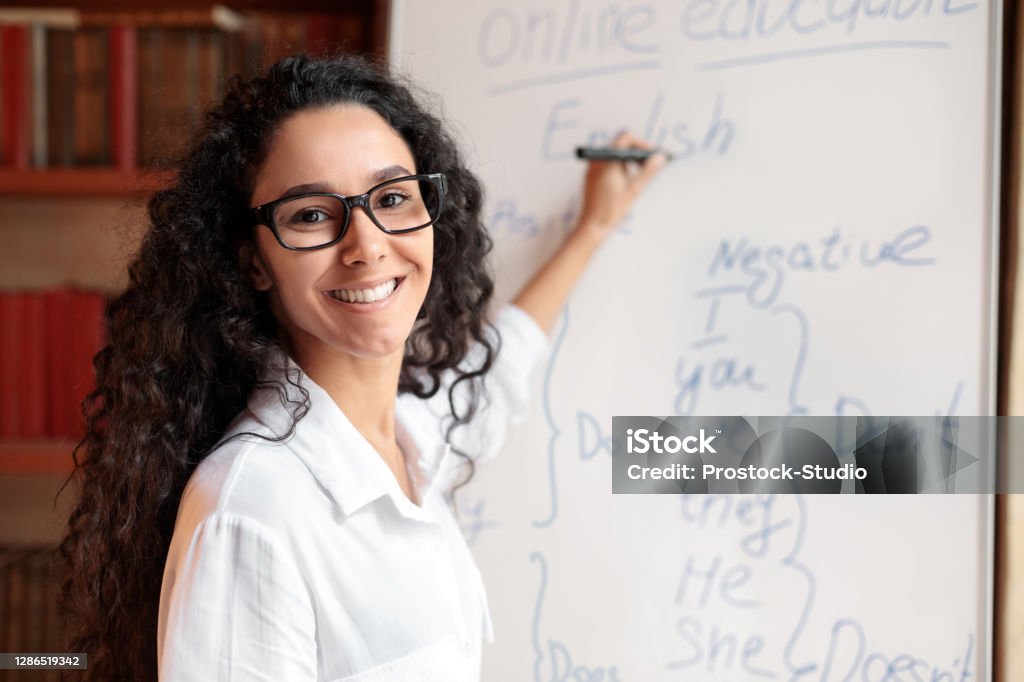 English teacher standing at board, explaining lesson to students Education And Learning Concept. Portrait of smiling female teacher standing at whiteboard, explaining grammar rules to students. Excited woman in glasses looking at camera, writing on the board Teacher Stock Photo