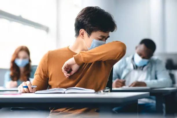 Photo of Asian male student couching in his elbow, sneezing in sleeve