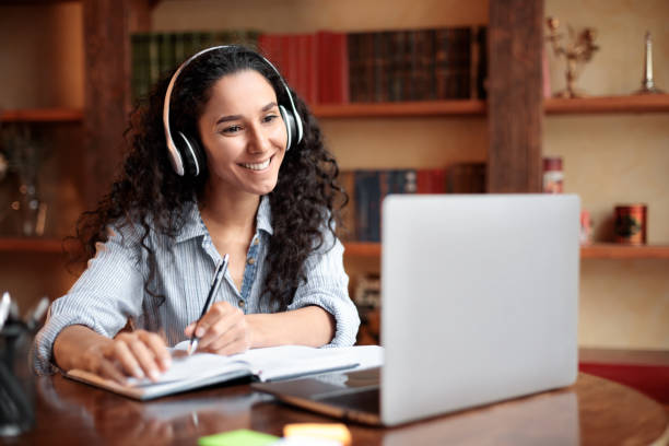 Woman sitting at desk, using computer and writing in notebook Home Office. Portrait of smiling young lady in wireless headphones sitting at desk, writing in notebook and using computer. Woman watching online tutorial, webinar or seminar, studying at home e learning stock pictures, royalty-free photos & images