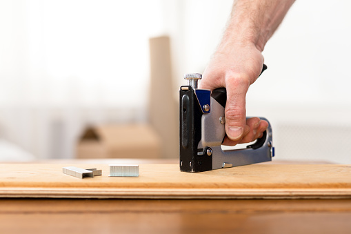Carpentry. Male Hand Using Staple Gun Tool, Unrecognizable Carpenter Man Working Making Furniture Using Professional Tools Indoor, Closeup, Cropped. Furniture Assembling And Repair Concept