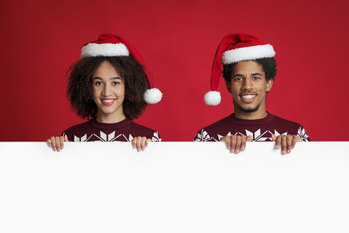 Winter mood and Christmas celebration. Cheerful young african american friends or family in Xmas sweaters and hats, holds white poster with empty space, isolated on red background, studio shot
