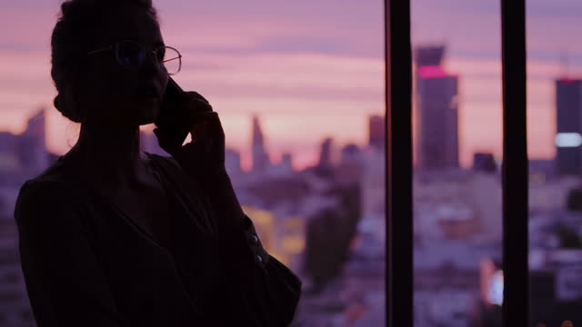 Sunset cityscape in office windows. Elegant businesswoman's silhouette making a call
