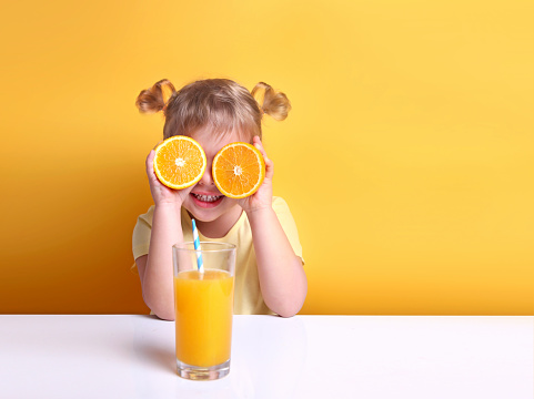 Child girl with oranges on eyes sitting at empty space table with glass of orange juice.Healthy vitamin child's nutrition.Kid with fruits.Funny toddler.