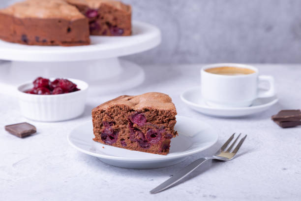 Chocolate cake with cherries on a white plate. In the background is a pie, a cup of coffee, cherries and pieces of chocolate.