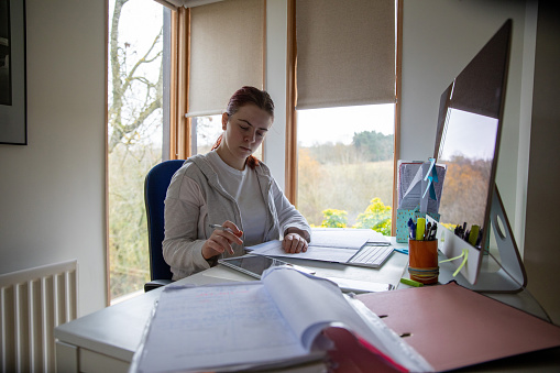 Student studying from home in an office during the Covid 29 pandemic. She is reading books, writing notes and is using a desktop computer.