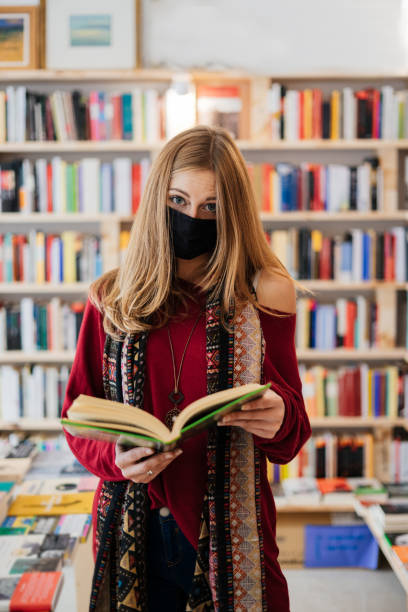 Beautiful blonde woman wearing a face mask with a book in a bookstore looking at the camera. stock photo