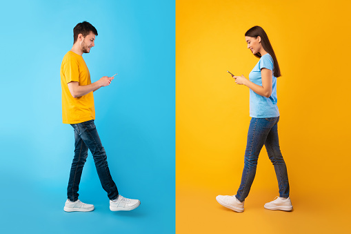 Communication, Technology And Lifestyle Concept. Full length portrait of smiling young couple using mobile phones, walking isolated over blue and yellow studio background, chatting