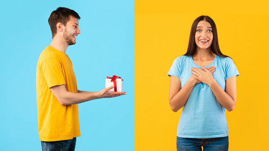 This Present Is For You. Portrait of smiling guy holding wrapped present box and giving it to his girlfriend, isolated over blue and yellow studio background, impressed woman holding hands on chest