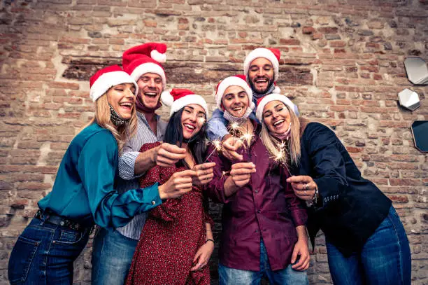 Group of happy friends with the face mask down in New Year eve with sparkling bengal lights - Group of young coworkers
celebrating Christmas wearing  Santa hats - Happy people in the close up