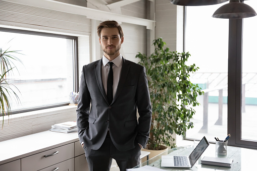 Portrait of smiling confident handsome young businessman in stylish formal suit standing in modern workplace. Successful ceo executive manager leader financial consultant lawyer looking at camera.