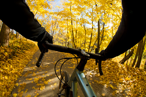 The handlebar view of a man riding his bicycle on a quiet, country road near Nelson, British Columbia, Canada in autumn. He is riding a gravel-style or cyclo-cross bicycle that is similar to a road bike but has larger tires which are more suited to rough terrain. The bicycle also has disc brakes.